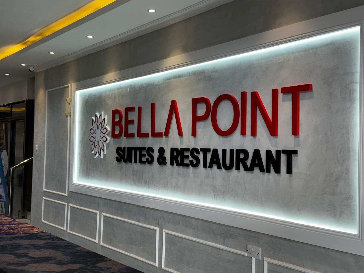 The illuminated sign in the Bellapoint Suites lobby, lettered "Bella Point Suites & Restaurant", on a grey polished-plaster wall.